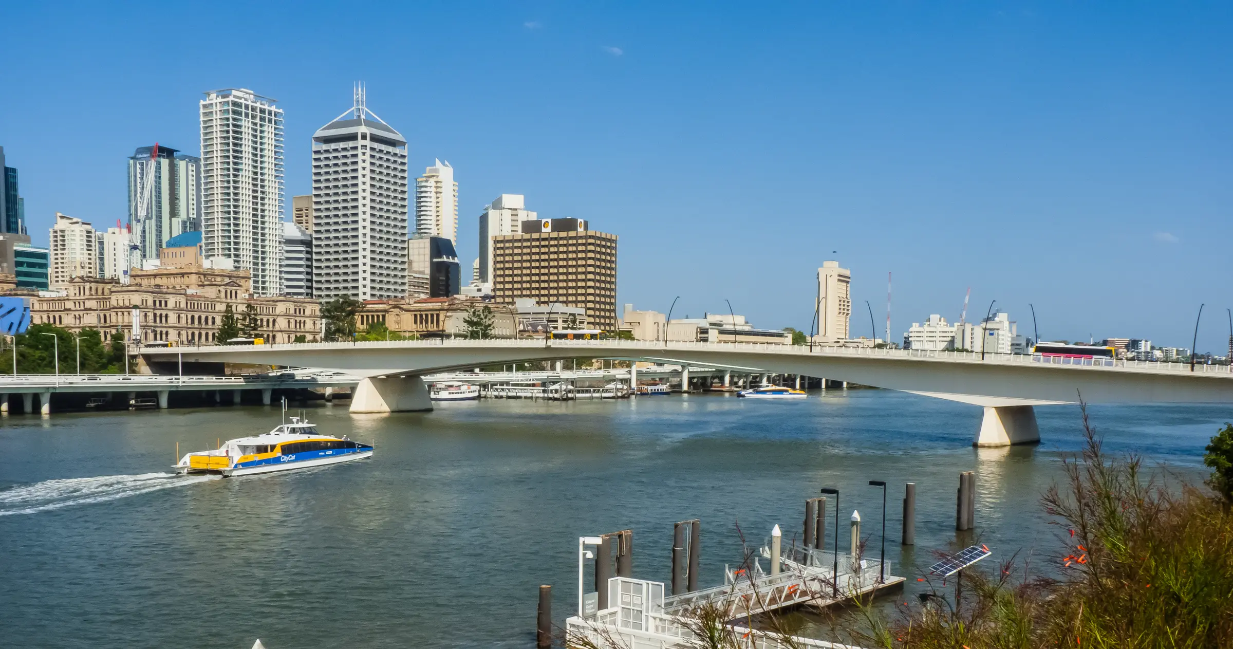A blue and yellow CityCat ferry crossing the Brisbane River towards the Victoria Bridge, sandstone buildings and the city towers behind under a clear blue sky