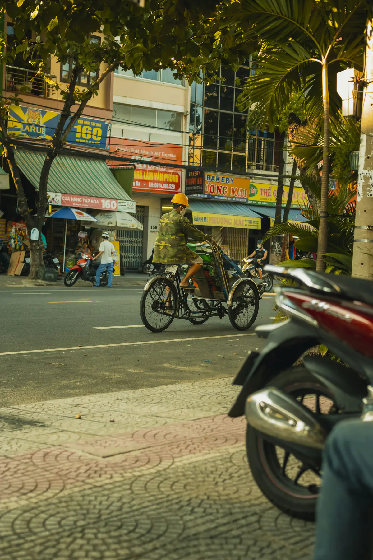 A cyclo driver in a yellow helmet pedals past bakery and shop signs on a Đà Nẵng street in late afternoon light