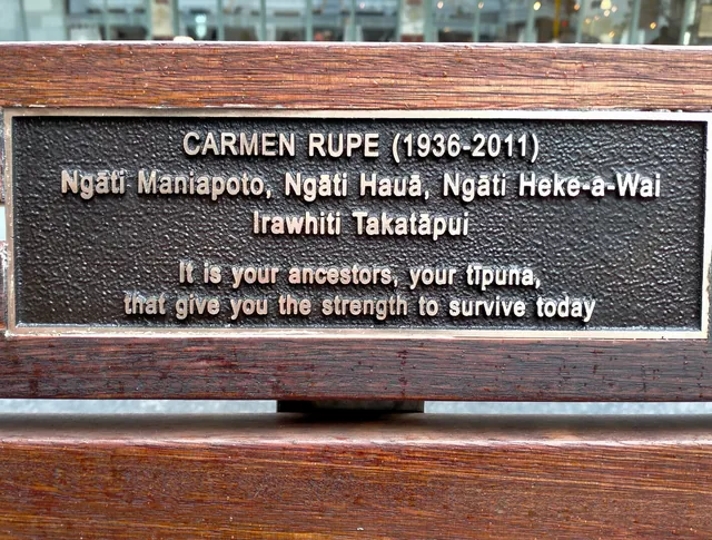A bronze plaque on a timber seat: Carmen Rupe, 1936 to 2011, her iwi, and the words: it is your ancestors, your tīpuna, that give you the strength to survive today